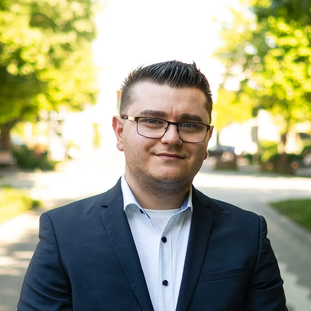 Ein Mann mit Brille im Anzug steht vor Fenstern mit Jalousien.
