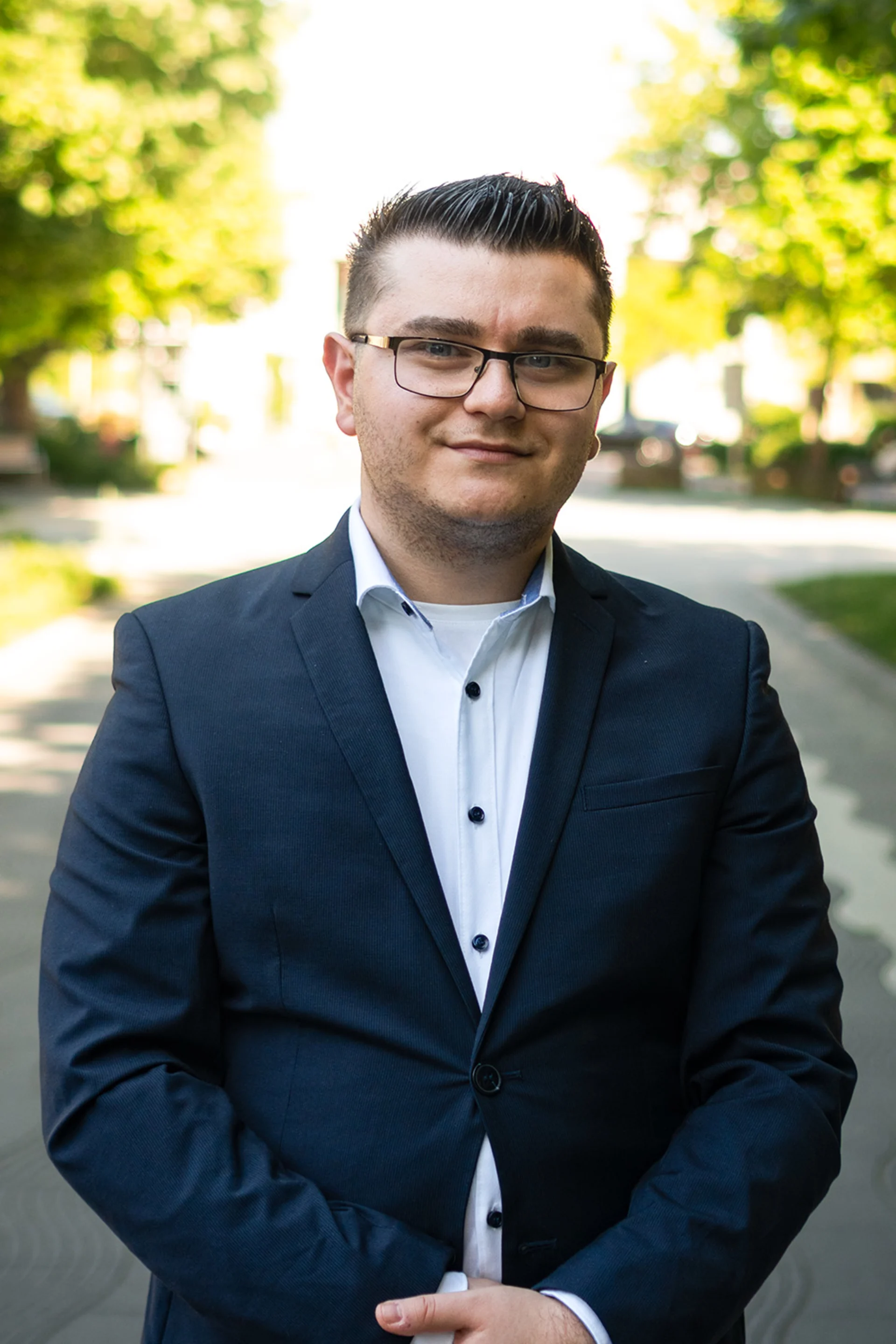Ein Mann mit Brille im Anzug steht vor Fenstern mit Jalousien.
