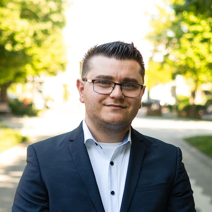 Ein Mann mit Brille im Anzug steht vor Fenstern mit Jalousien.