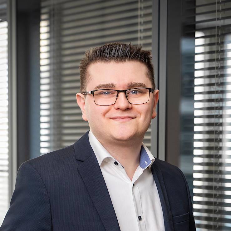 Ein Mann mit Brille im Anzug steht vor Fenstern mit Jalousien.
