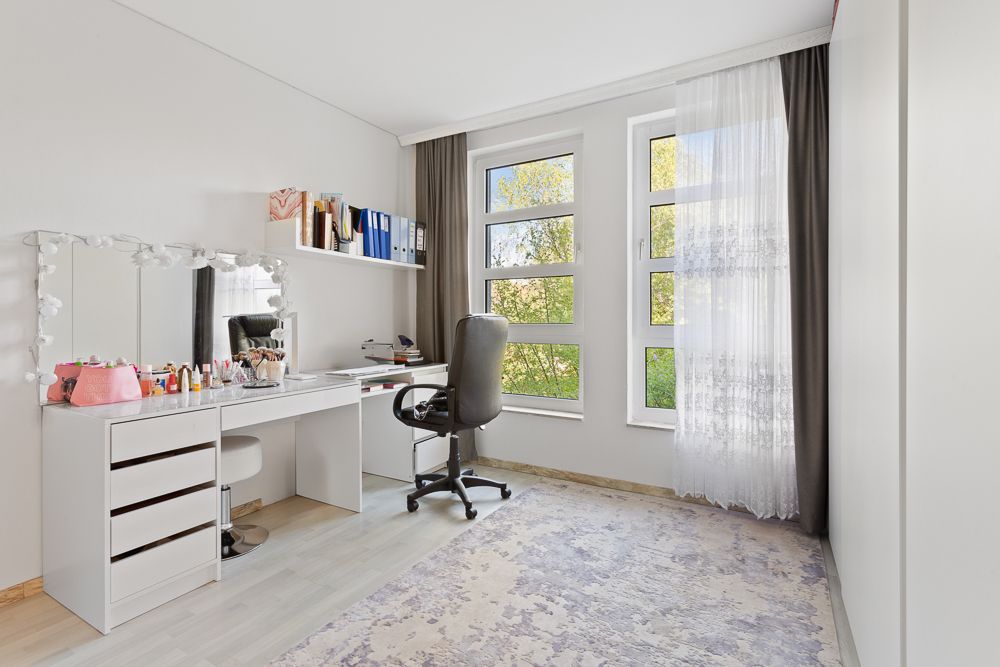 Helles Zimmer mit großem Fenster, Schminktisch, Stuhl und Regal mit Büchern und Ordnern.