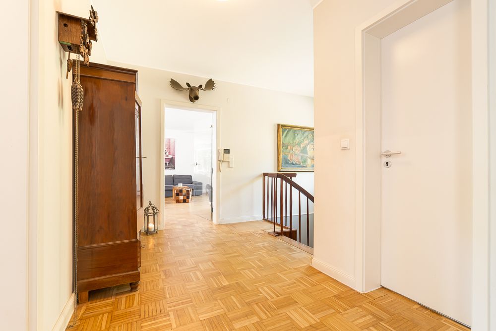 Heller Flur mit Parkettboden, Holzschrank, Treppengeländer und offener Tür zum Wohnzimmer.