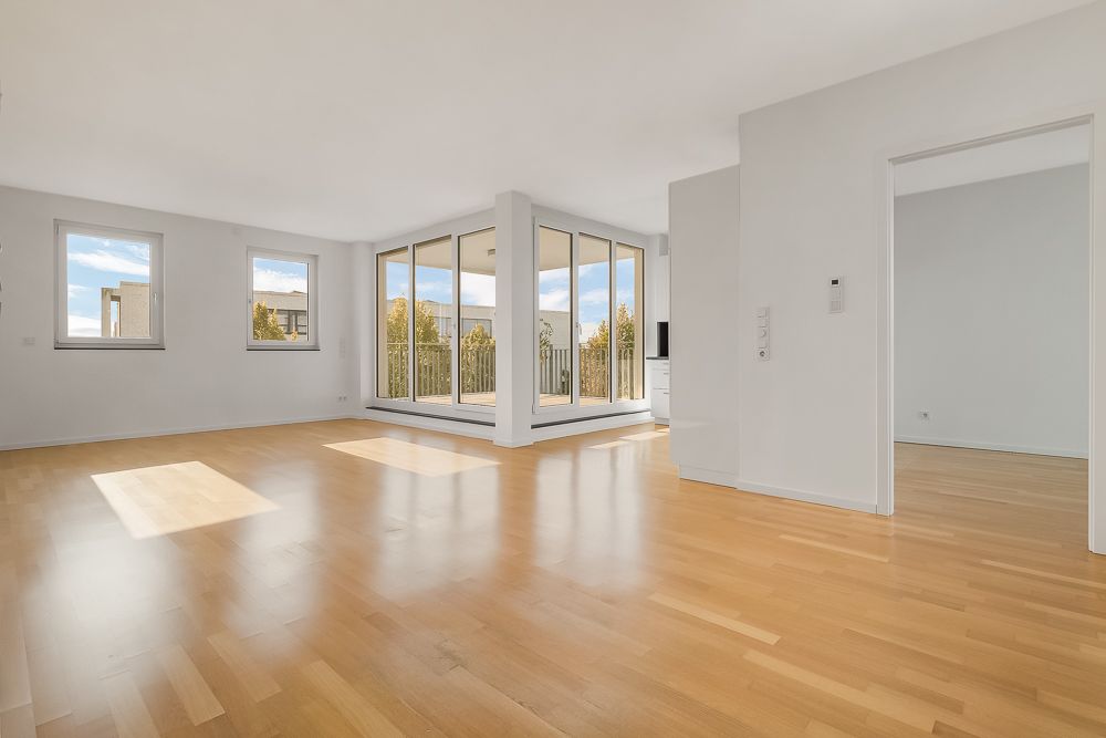 Heller Wohnraum mit Parkettboden, großen Fenstern und Zugang zu einem verglasten Balkon.