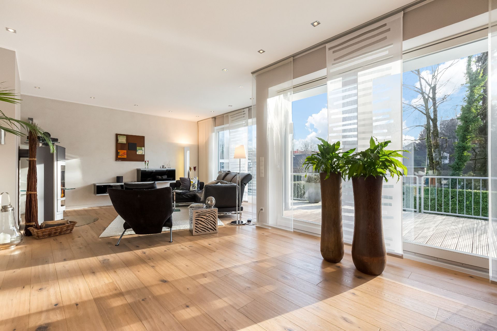 Helles Wohnzimmer mit Holzboden, großen Fenstern, zwei Pflanzen und modernem Mobiliar.