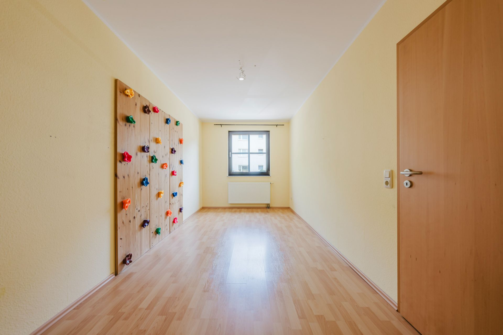 Heller Raum mit Holzboden, Fenster und Kletterwand an der linken Wand.