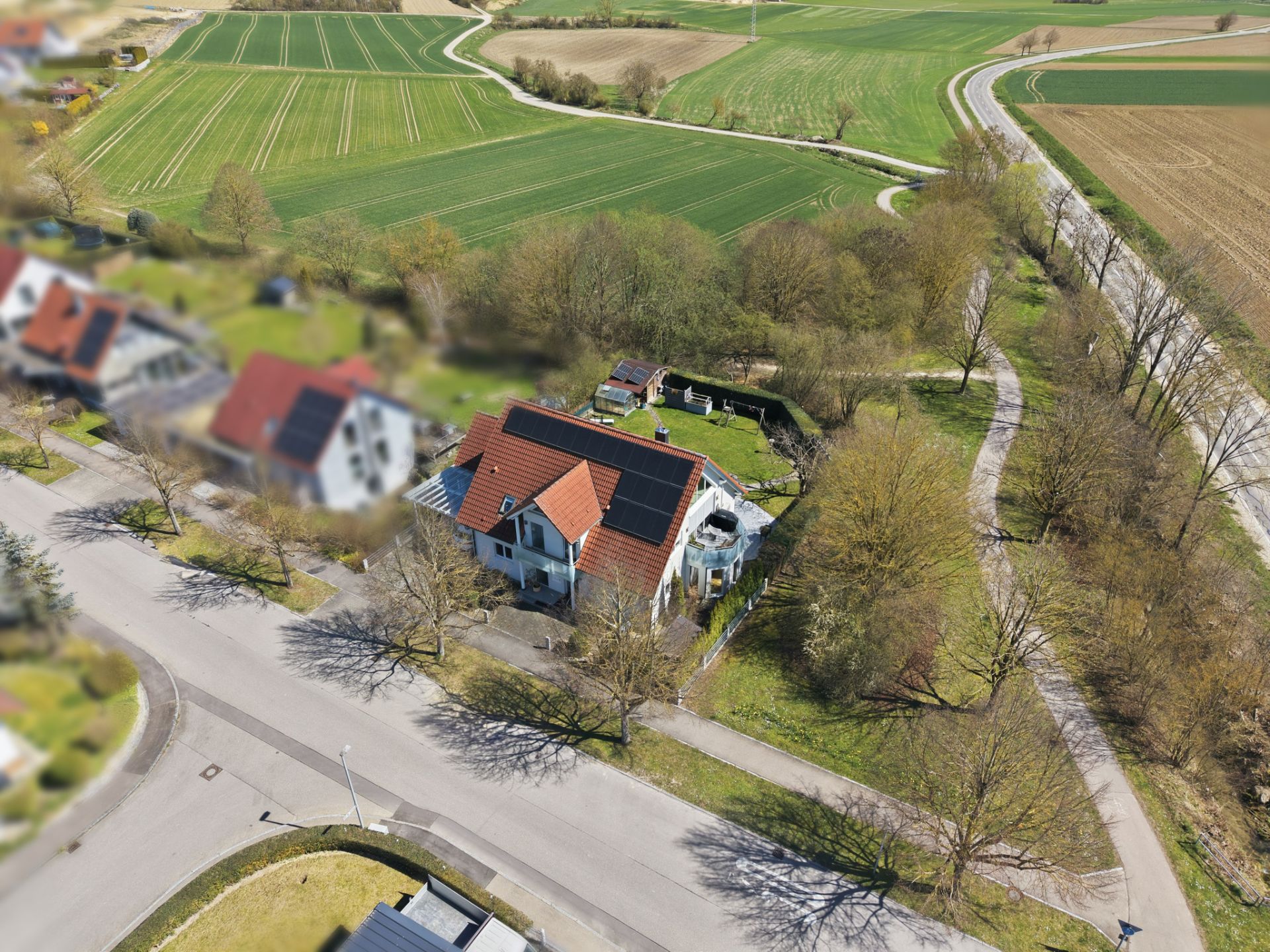 Luftaufnahme eines Einfamilienhauses mit Garten an einer ruhigen Straße, umgeben von Feldern und Bäumen.