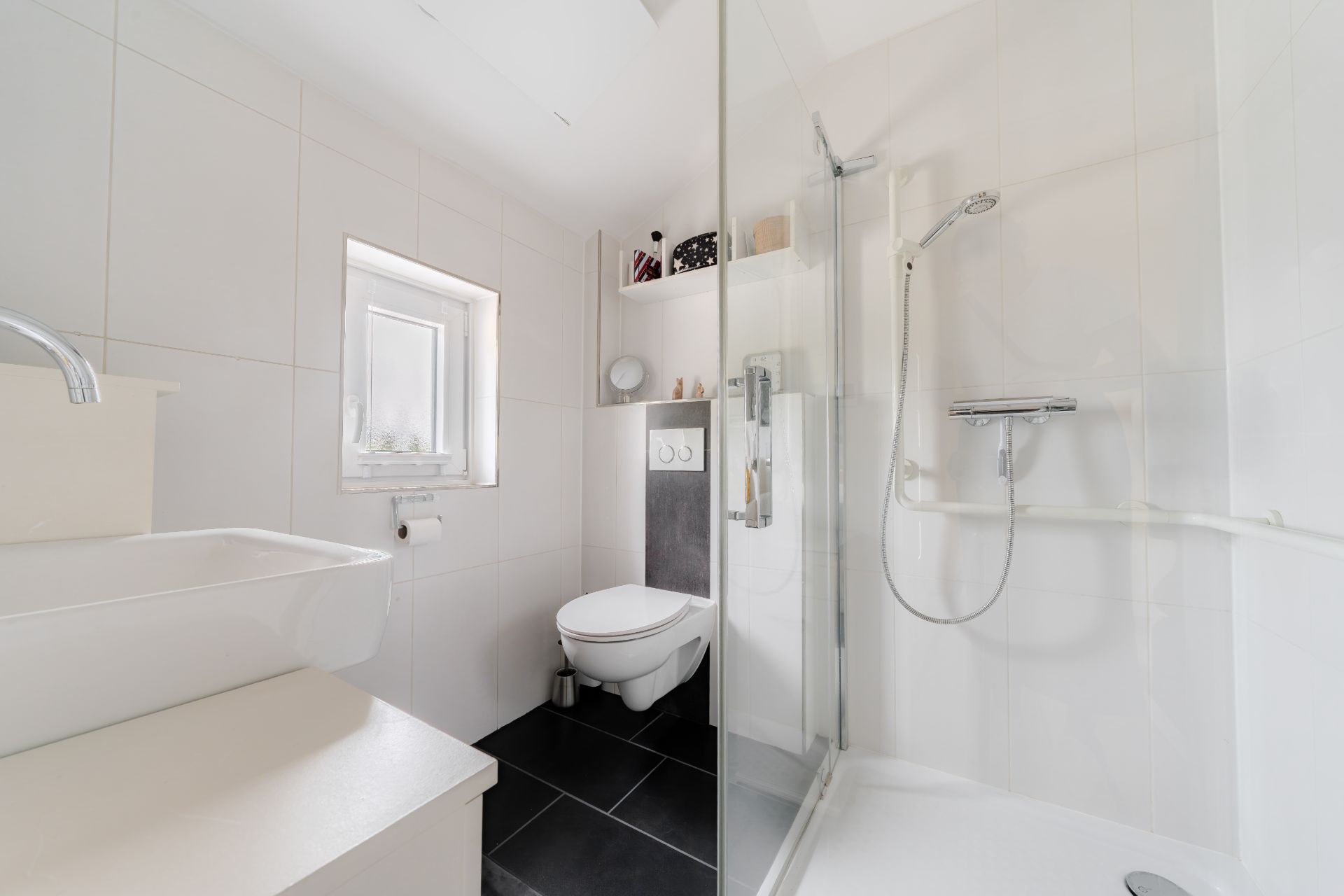 Helles Badezimmer mit Waschbecken, Toilette, Fenster und Dusche mit Glaswand.