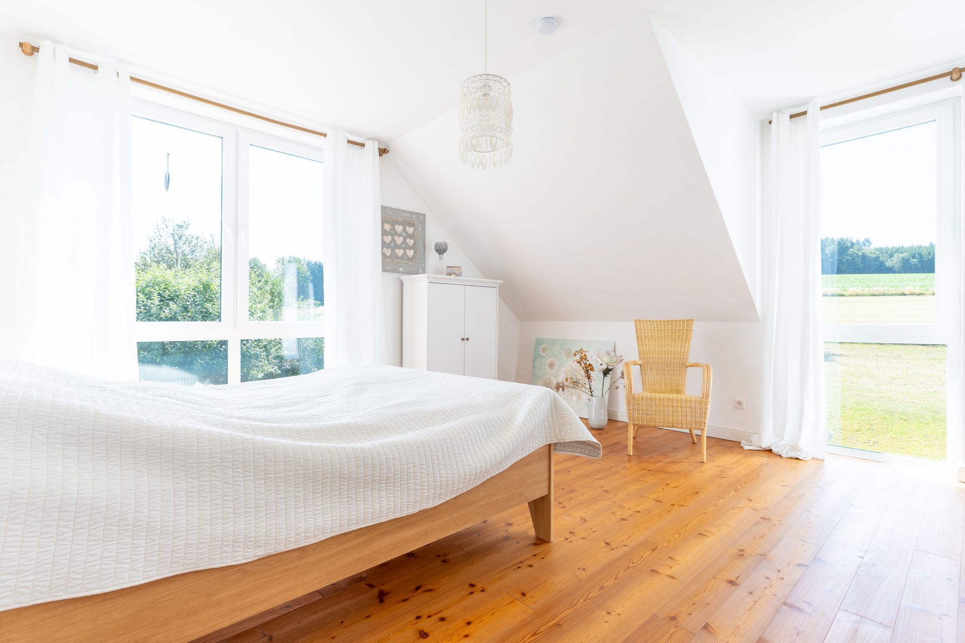 Helles Schlafzimmer mit Holzboden, Bett, Stuhl, Schrank und großen Fenstern mit weißem Vorhang.