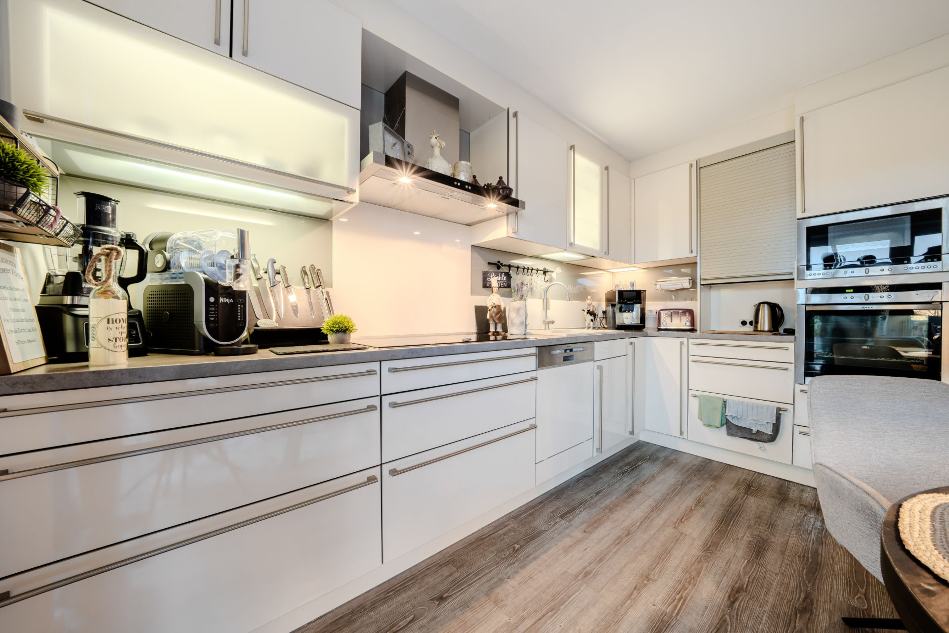 Moderne weiße Küche mit Einbaugeräten, Holzfußboden und beleuchteten Hängeschränken.