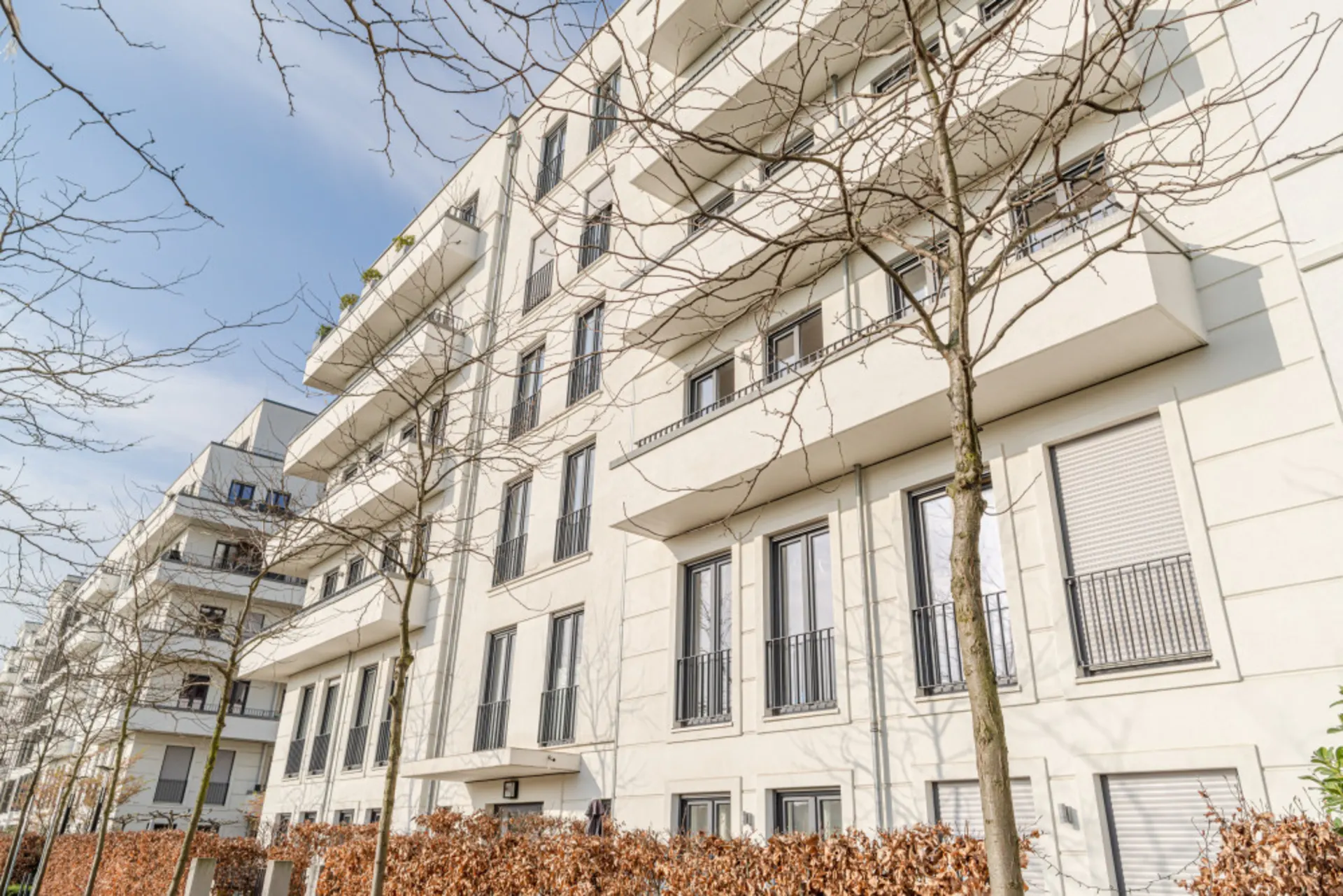 Helles Wohnhaus mit mehreren Balkonen und kahlen Bäumen im Vordergrund.