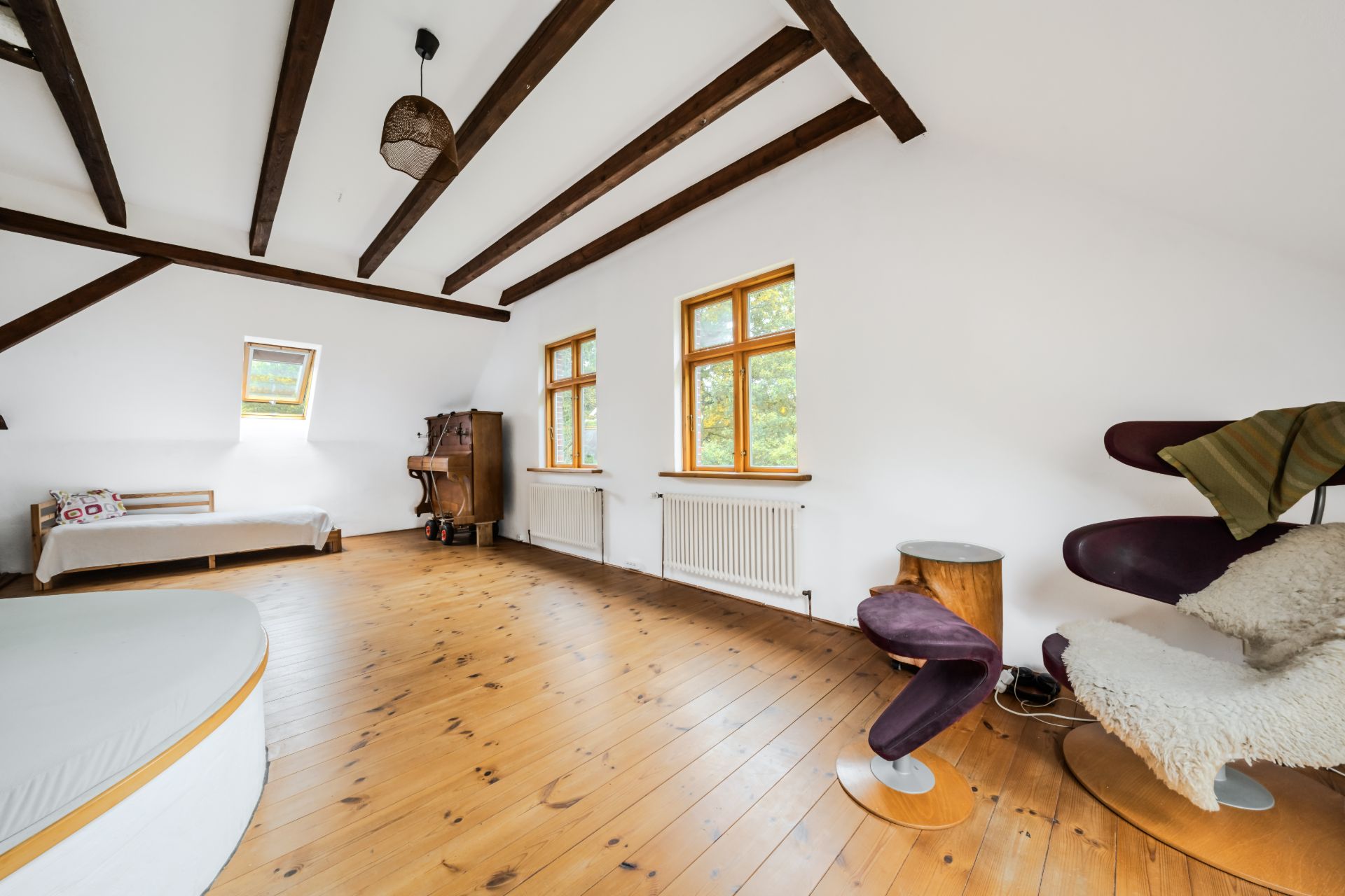 Helles Zimmer mit Holzboden, sichtbaren Deckenbalken, Sessel, Bett und großen Fenstern.