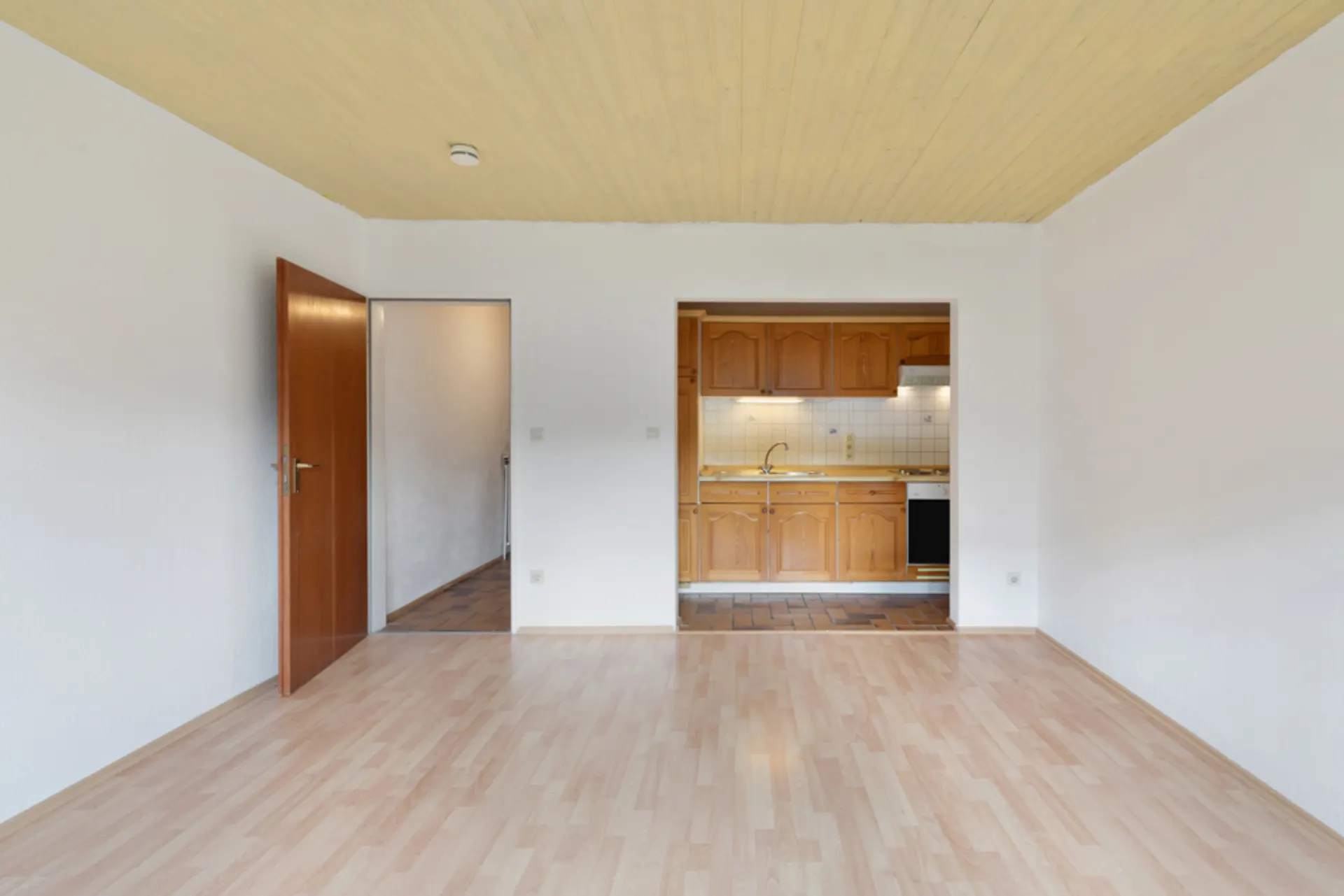 Leeres Zimmer mit Holzboden, offener Tür und Einbauküche im Hintergrund.