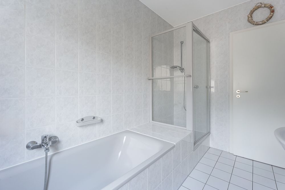 Helles Badezimmer mit Badewanne, Dusche mit Glastür und weiß gefliesten Wänden und Boden.