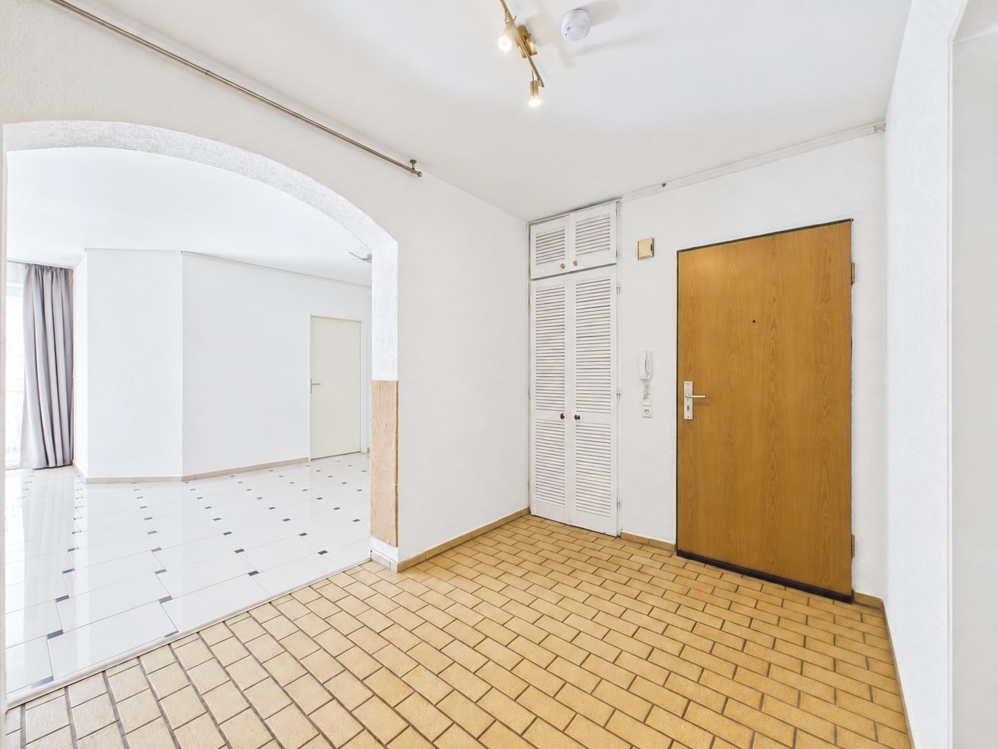 Heller Flur mit Holztür, weißen Wänden und Durchgang zum angrenzenden Raum mit Fliesenboden.