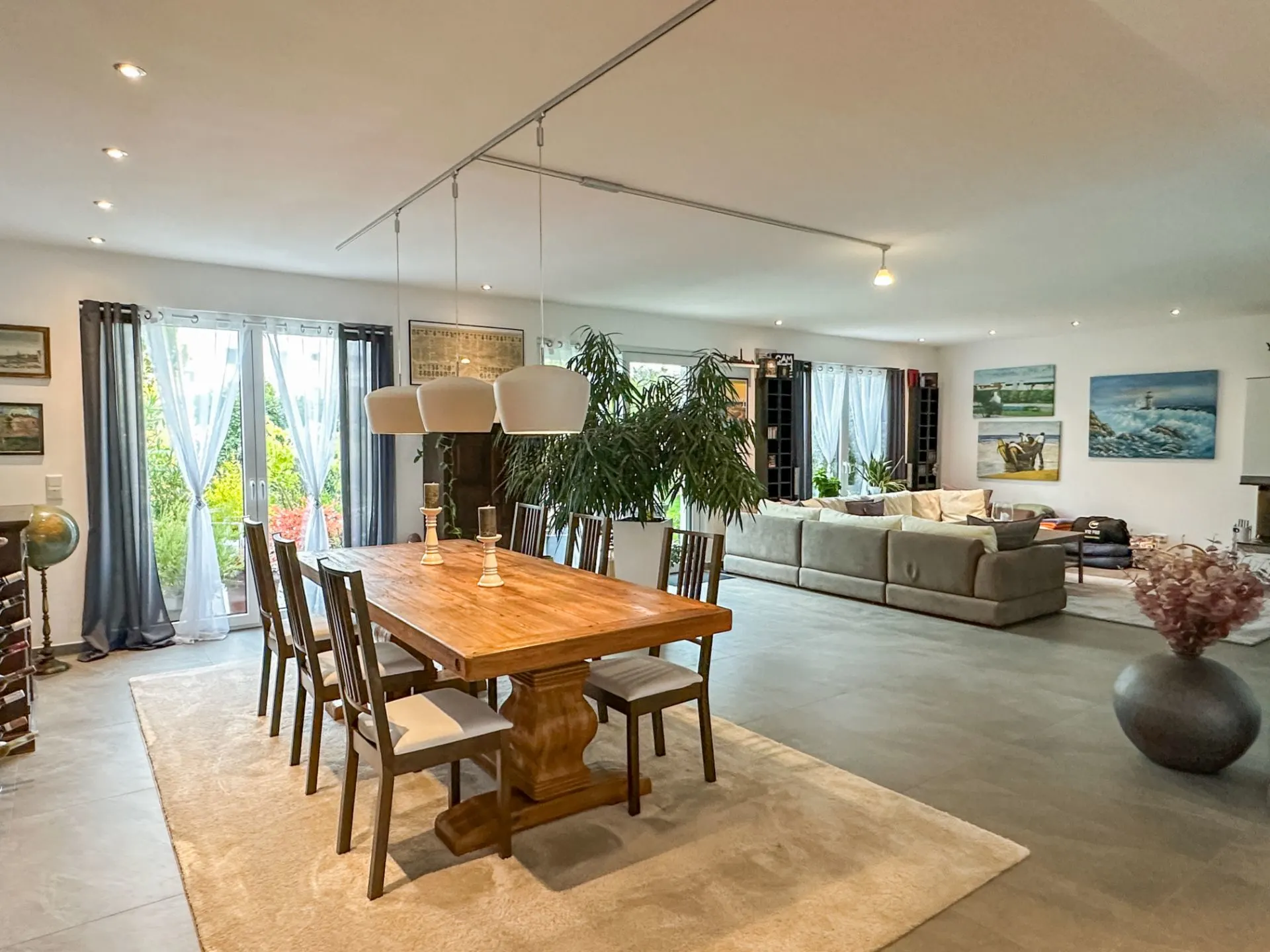 Helles Wohnzimmer mit großem Esstisch, Stühlen, Sofa, Pflanzen und großem Fenster.
