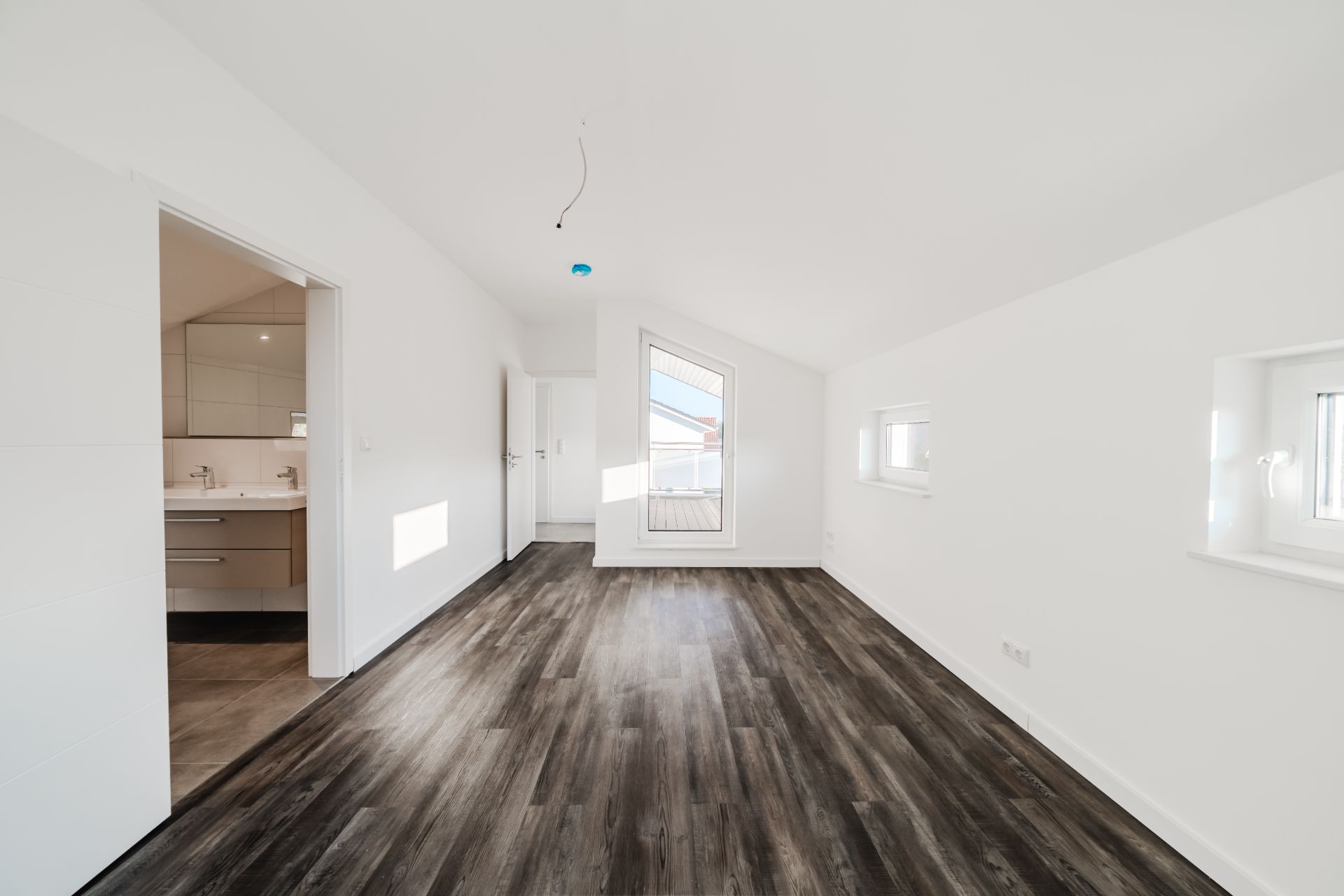 Leerer, heller Raum mit dunklem Holzboden und weißen Wänden, Fenster und Tür zum Badezimmer.
