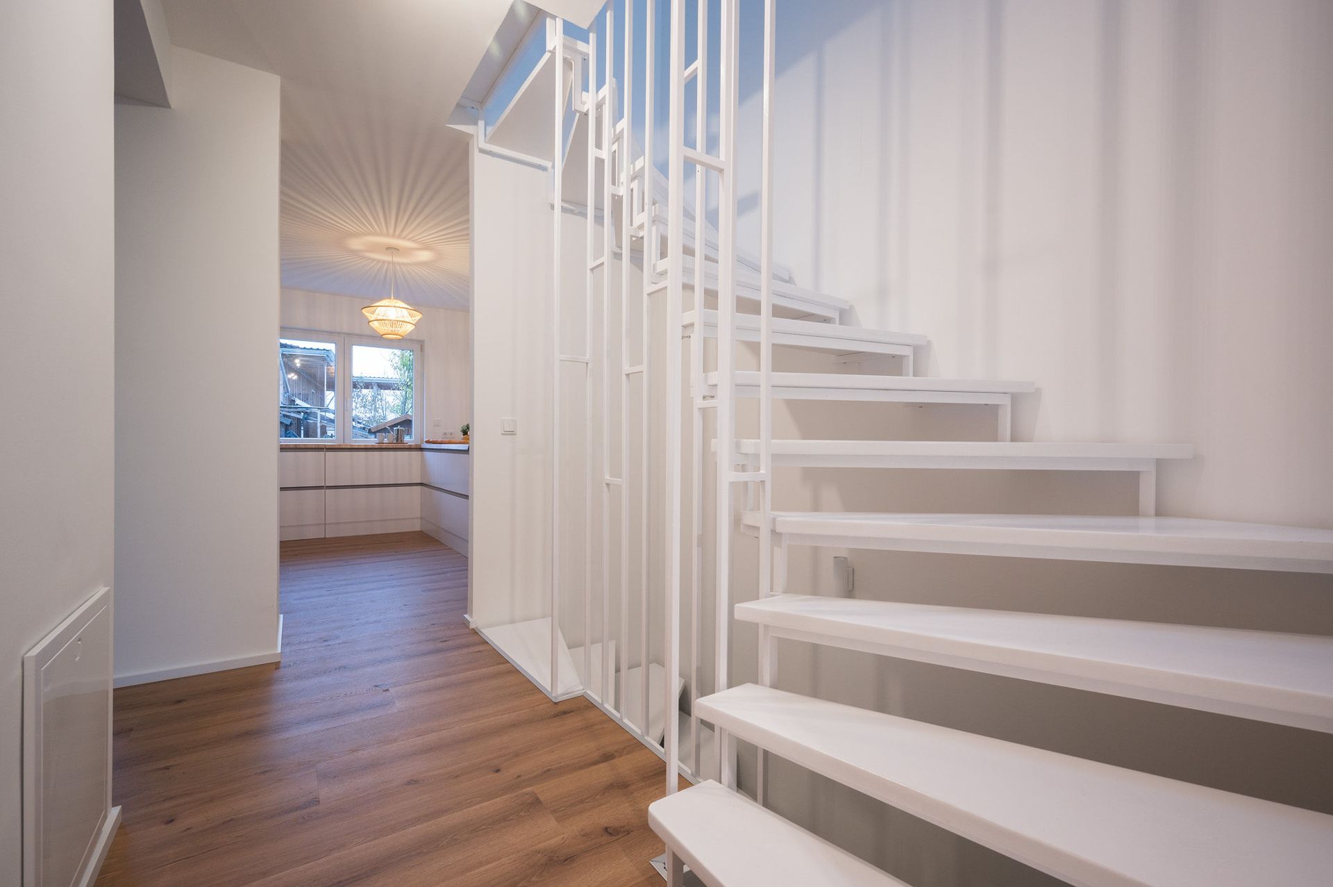 Weiße Treppe mit Geländer, daneben Flur mit Holzfußboden und Blick in eine helle Küche.