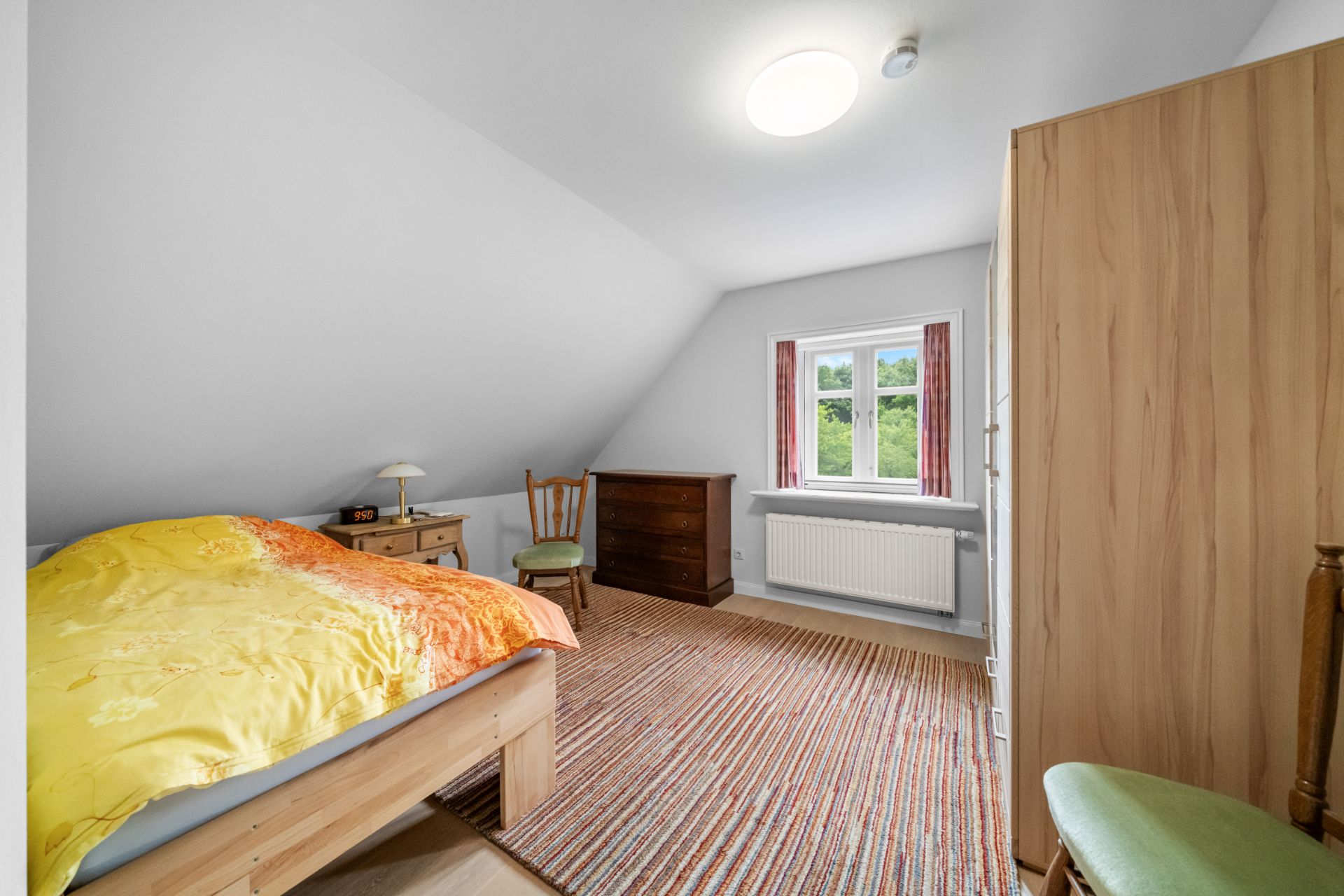 Helles Schlafzimmer mit Holzbett, Teppich, Schrank, Kommode und Fenster.