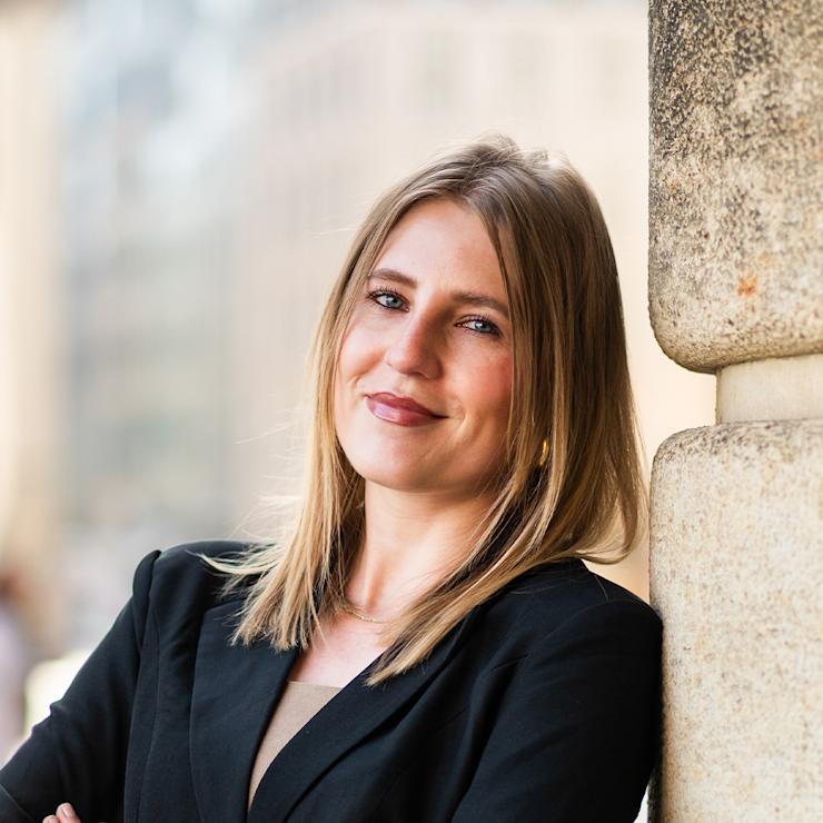 Junge Frau mit blondem Haar steht draußen an einer Steinwand und lächelt in die Kamera.