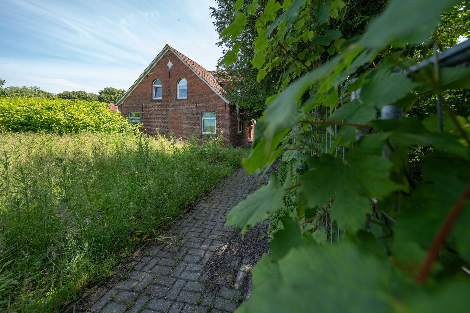 Backsteinhaus mit spitzem Dach, Weg und überwachsenem Garten im Vordergrund.