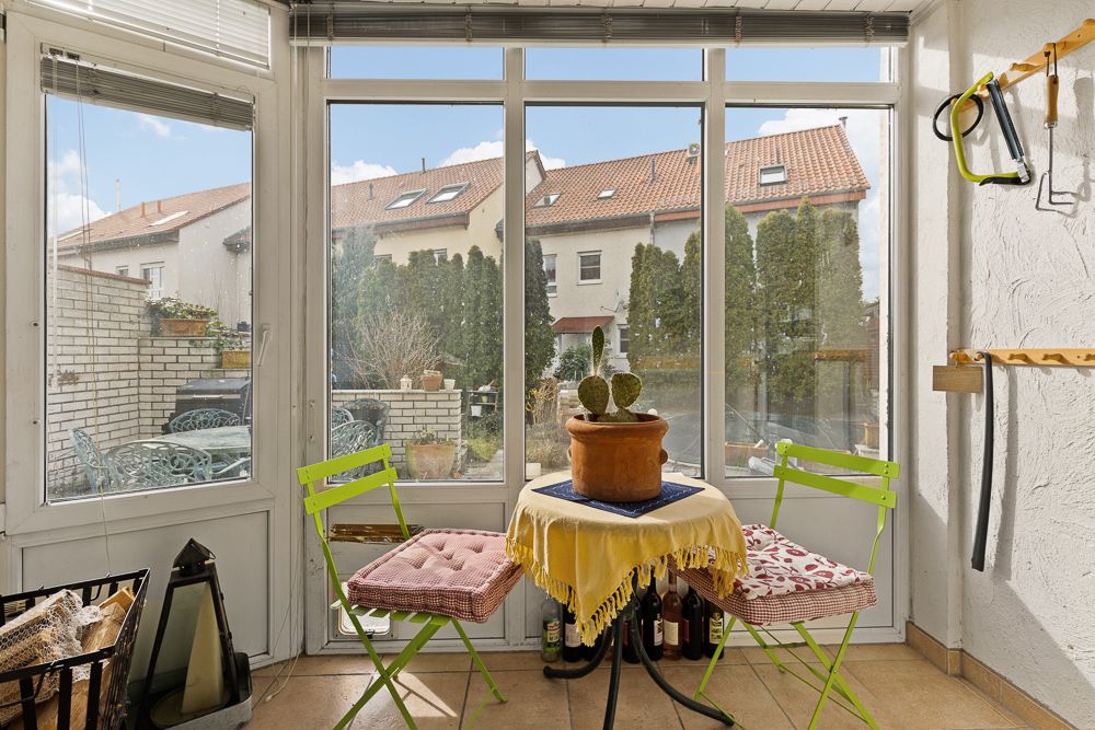 Wintergarten mit zwei grünen Stühlen, rundem Tisch und Kaktus-Pflanze, Blick auf Häuser und Garten.