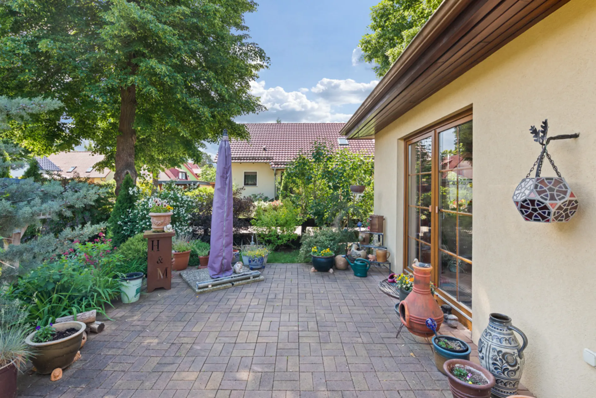 Gepflasterte Terrasse mit vielen Blumentöpfen und Gartenmöbeln neben einem Wohnhaus.