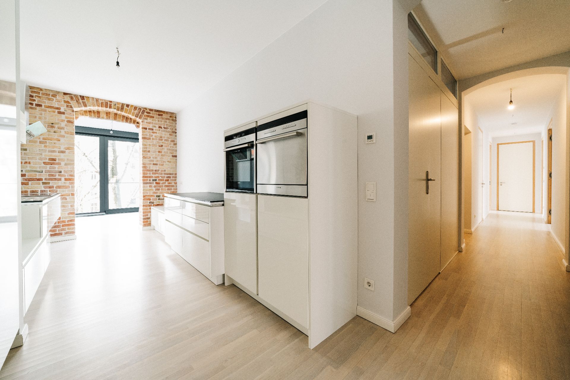 Helle moderne Küche mit weißen Schränken, Backofen und offenem Backsteinbogen, angrenzender Flur.