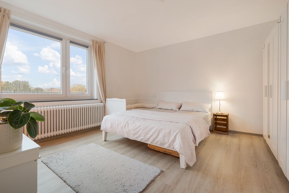 Helles Schlafzimmer mit Doppelbett, großem Fenster, Kleiderschrank und Nachttischlampe.