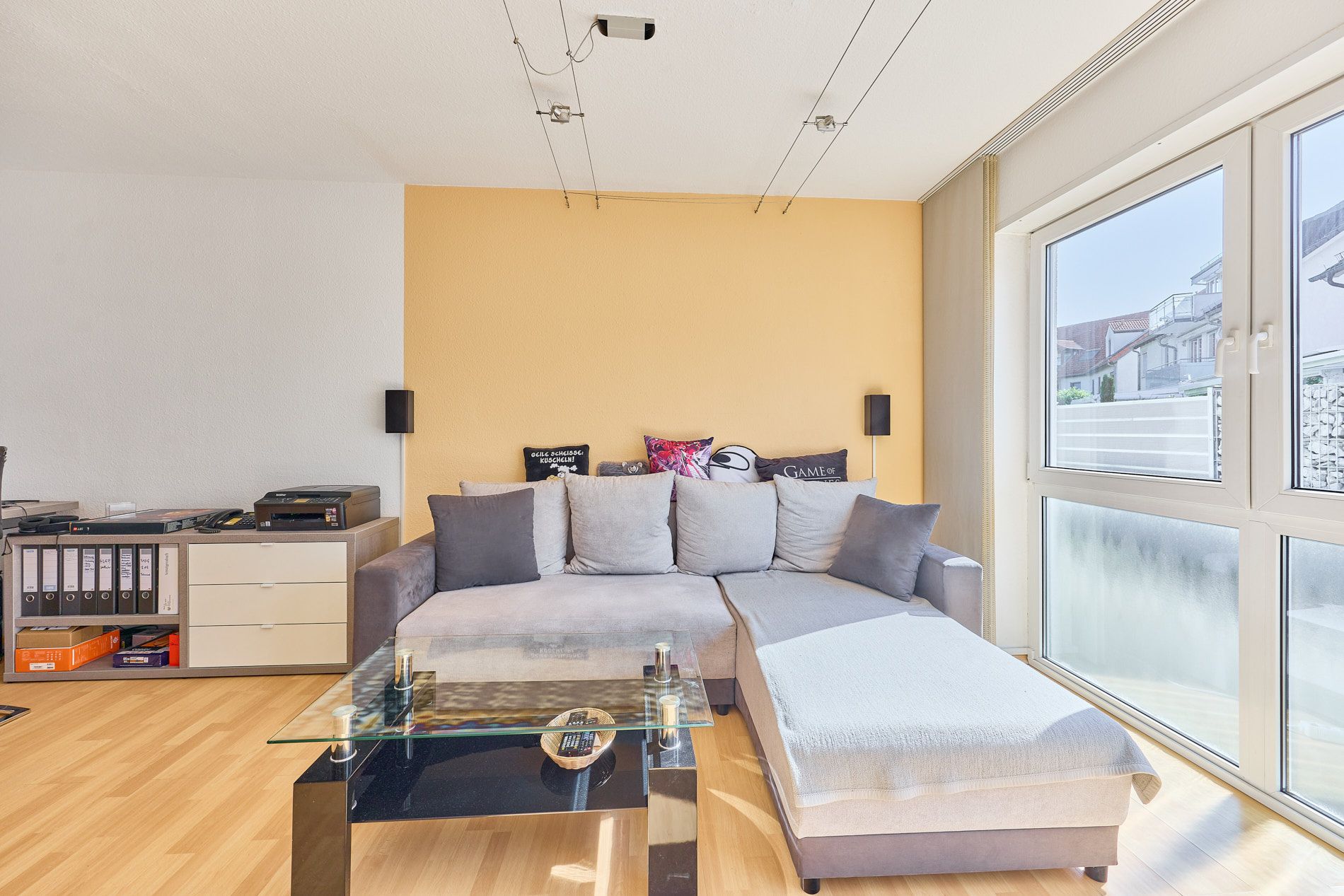 Helles Wohnzimmer mit Ecksofa, Glastisch und großem Fenster.