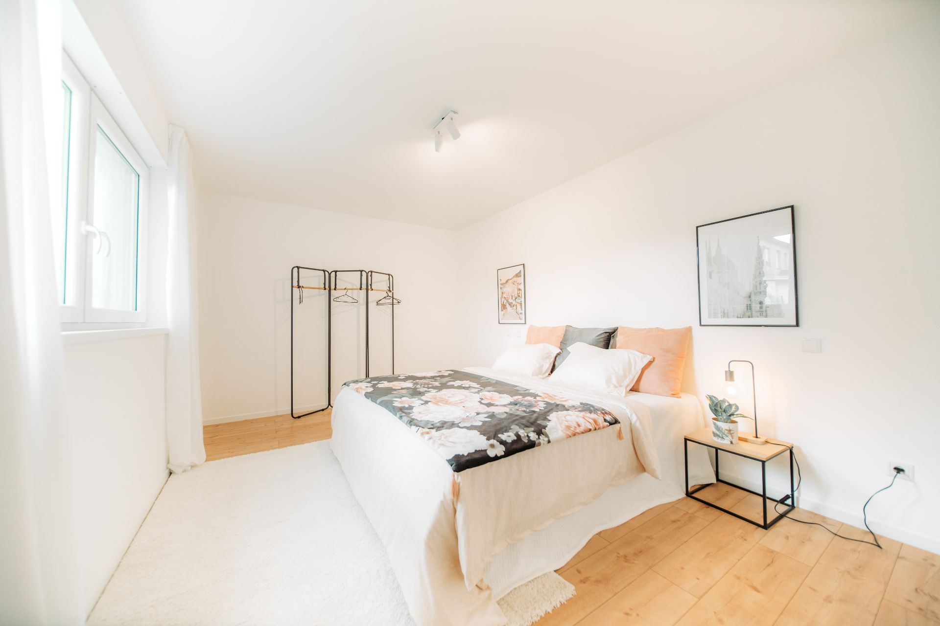 Helles Schlafzimmer mit Doppelbett, Nachttisch, offener Kleiderstange und Bildern an der Wand.