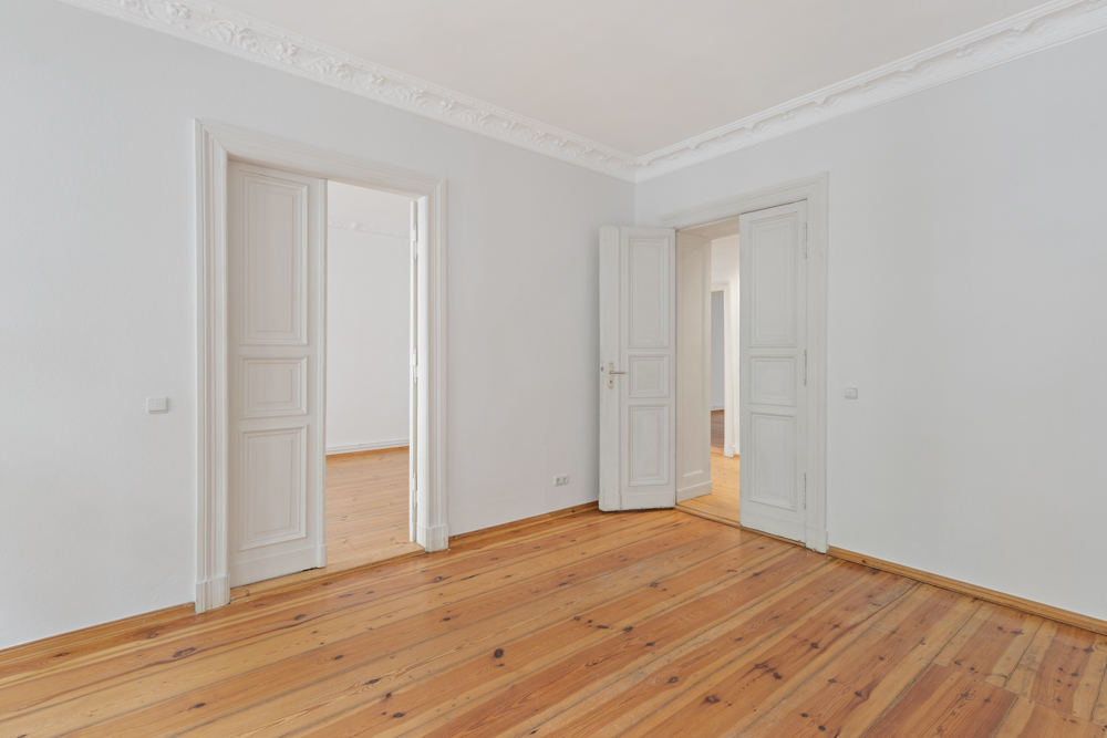Leerer Raum mit Holzboden, weißen Wänden und zwei offenen weißen Doppeltüren.
