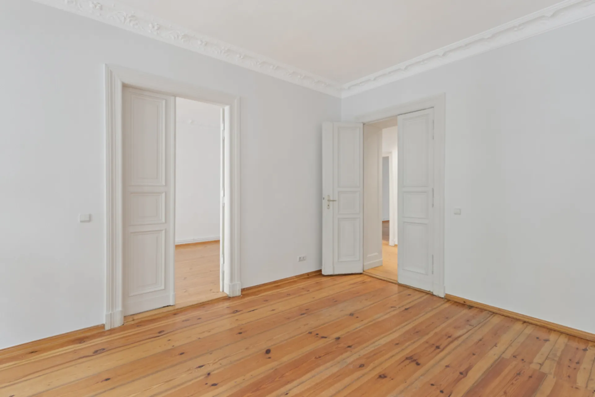 Leerer Raum mit Holzboden, weißen Wänden und zwei offenen weißen Doppeltüren.