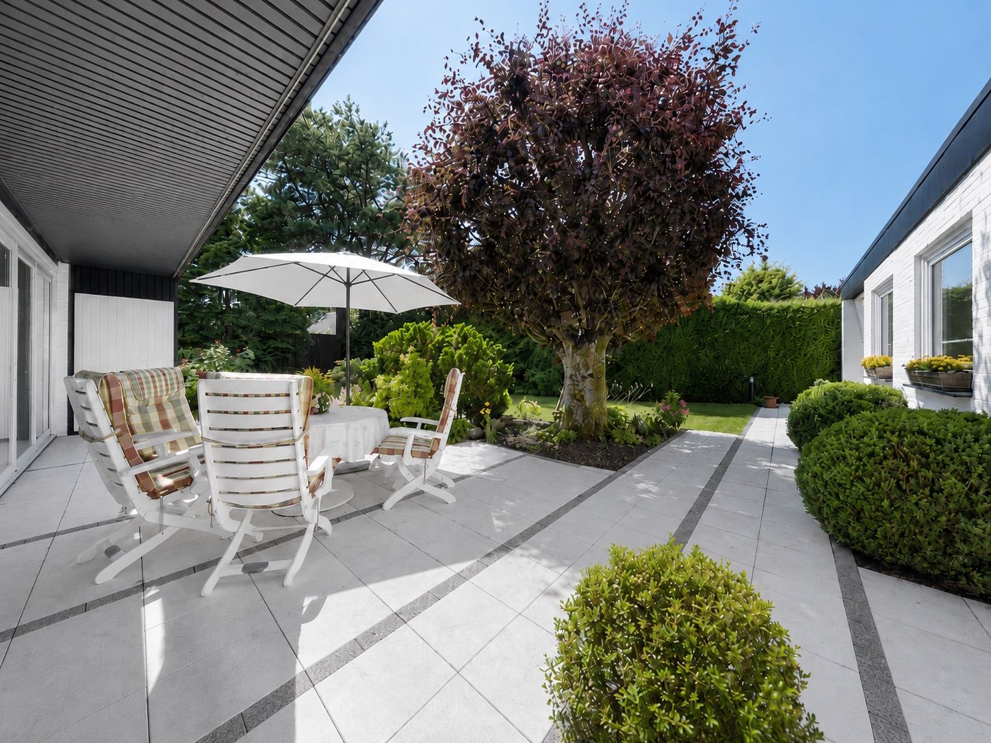Moderne Terrasse mit weißen Gartenmöbeln, Sonnenschirm und großem Baum im Garten.