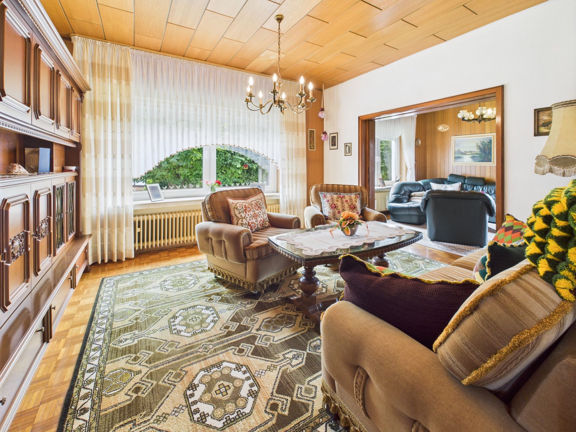 Gemütliches Wohnzimmer mit Teppich, Sofas, großem Fenster und Holzmöbeln, angrenzend an weiteres Zimmer.