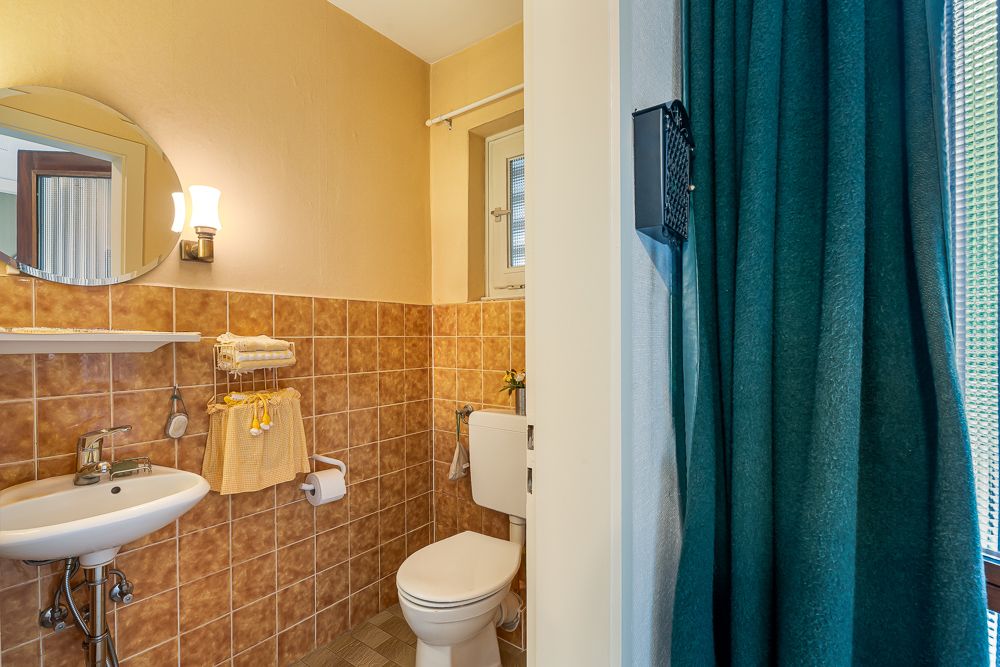 Kleines Badezimmer mit Toilette, Waschbecken und braunen Fliesen, Fenster und grünem Vorhang.