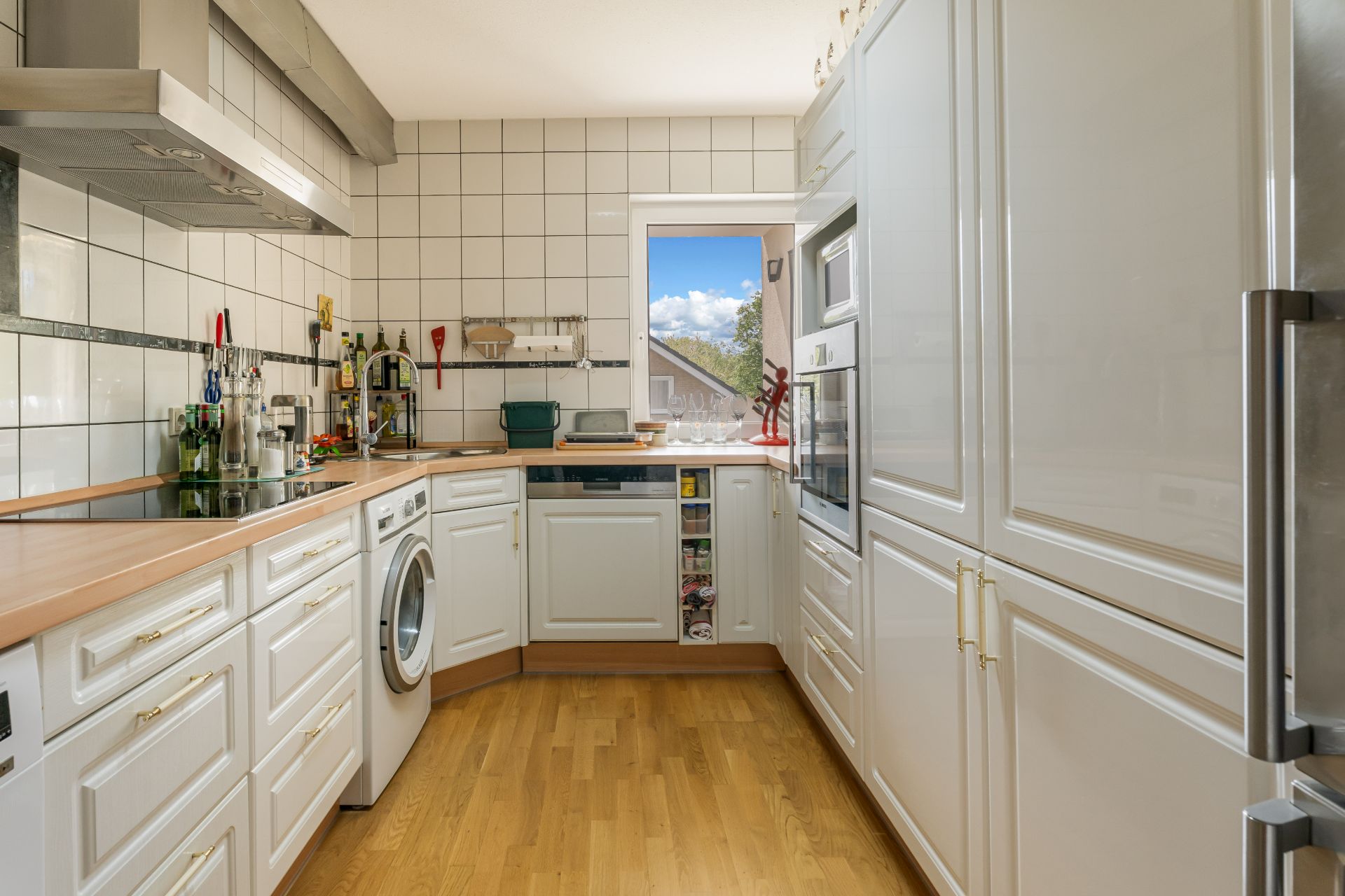 Moderne Küche mit weißen Schränken, Holzboden, Waschmaschine und Fenster mit Ausblick.
