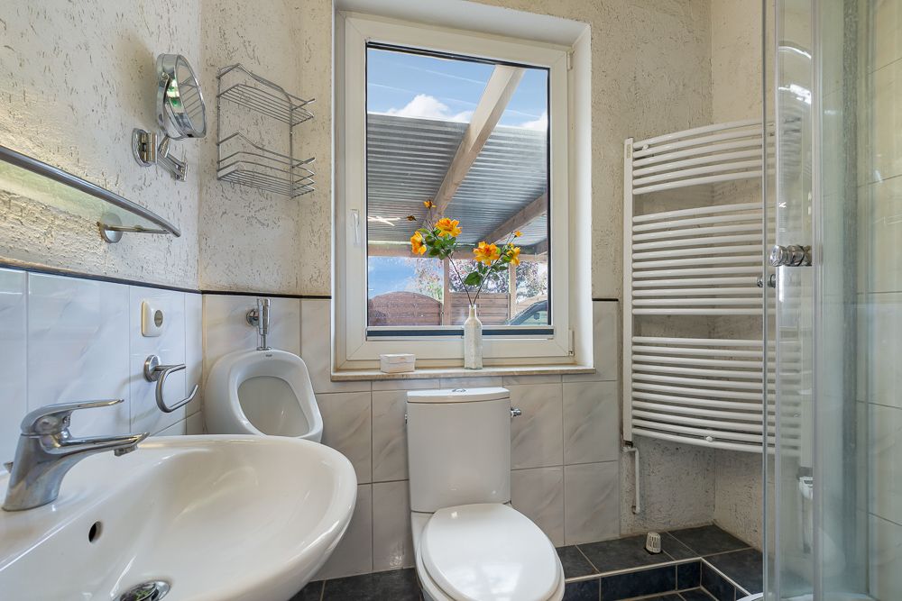 Modernes Badezimmer mit Waschbecken, Toilette, Urinal, Dusche, Heizkörper und Fenster mit Blumen.