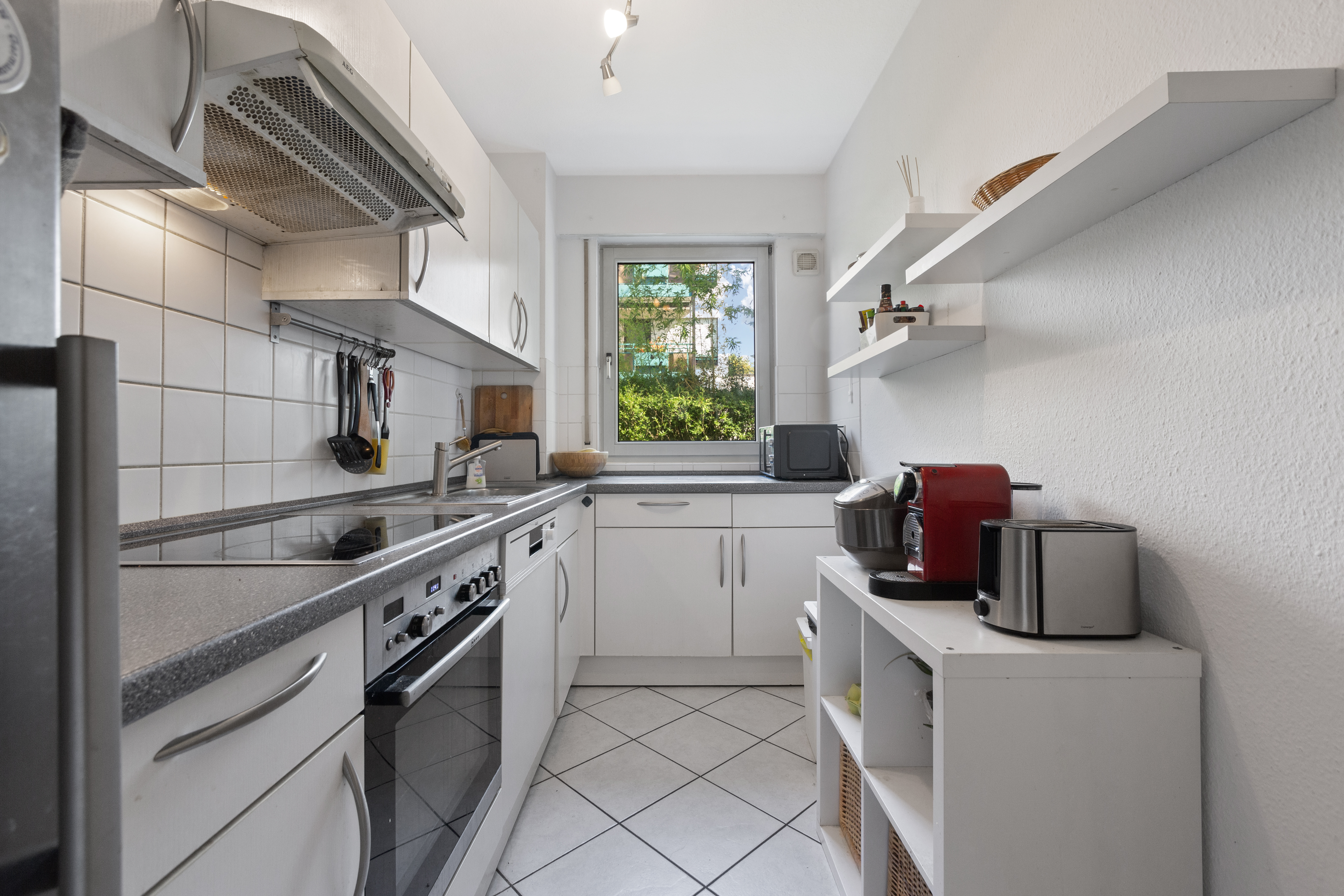 Moderne, schmale Küche mit weißen Schränken, Geräten und Fenster mit Blick ins Grüne.