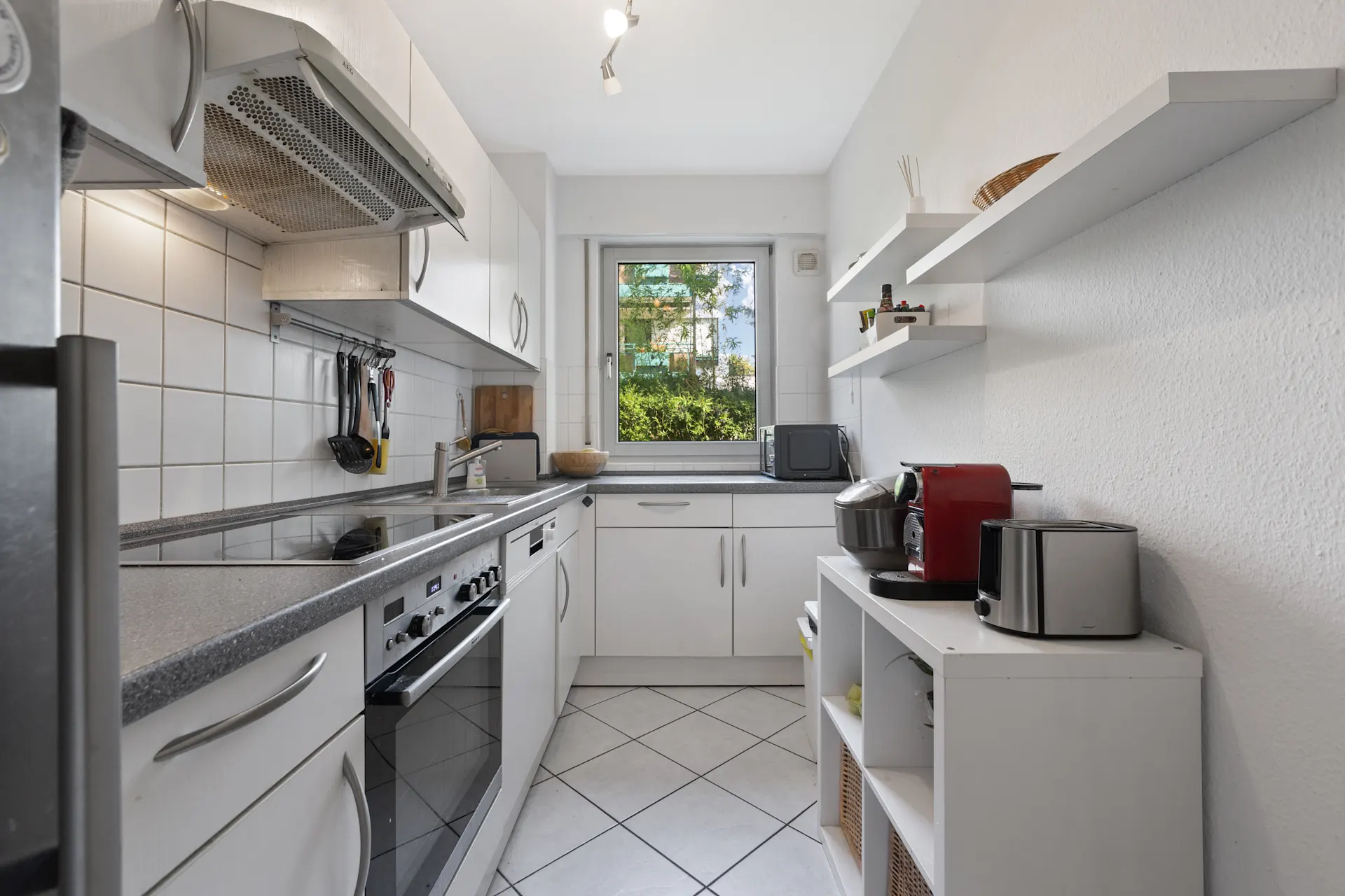 Moderne, schmale Küche mit weißen Schränken, Geräten und Fenster mit Blick ins Grüne.