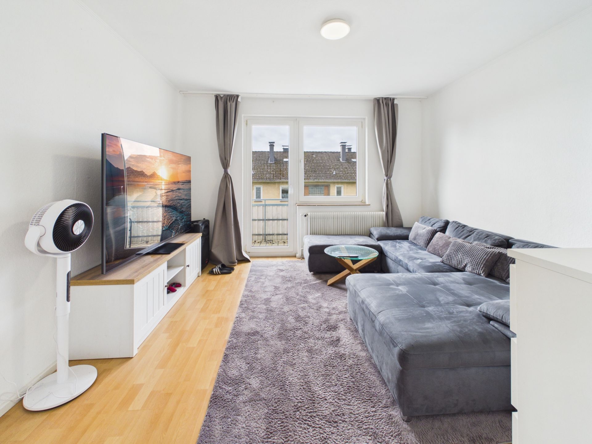 Wohnzimmer mit großer Ecksofa, Teppich, Fernseher, Stehventilator und Fenstertür mit grauen Vorhängen.