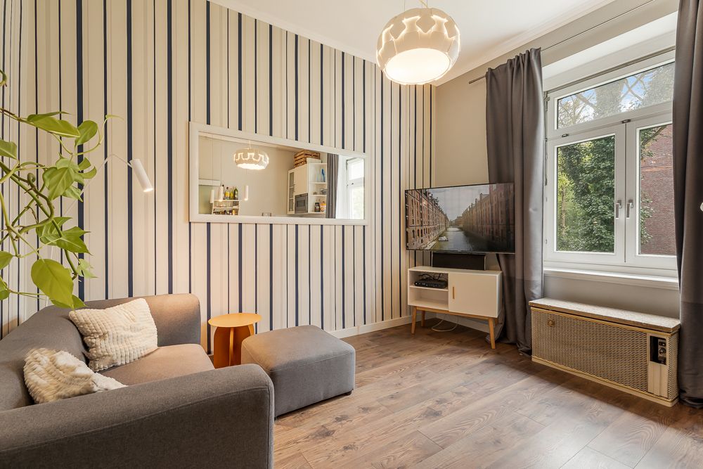 Modernes Wohnzimmer mit grauem Sofa, gestreifter Wand, Fernseher und großem Fenster mit Vorhängen.