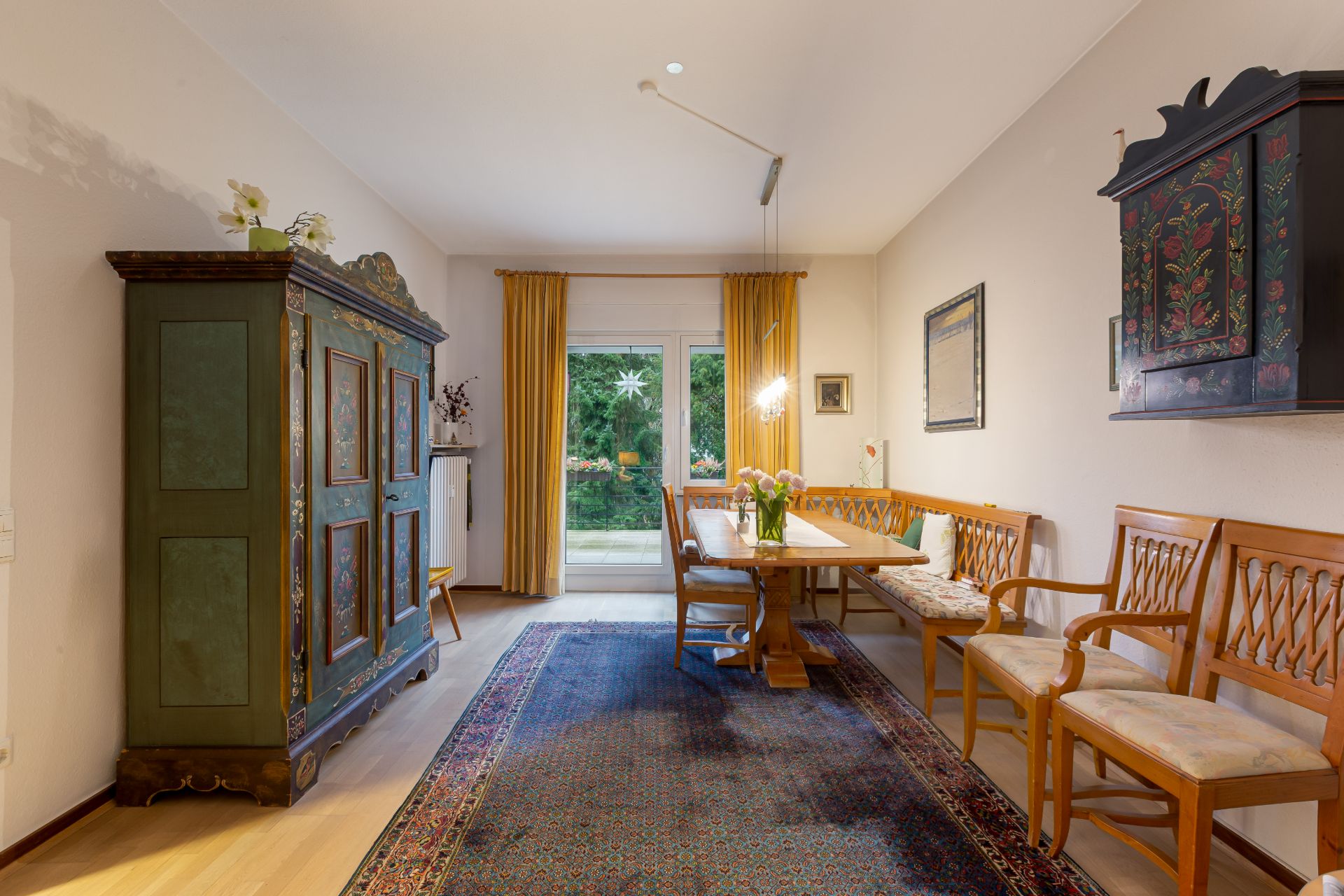 Wohnzimmer mit großem Teppich, Holzmöbeln, grünem Schrank und Blick auf den Garten.