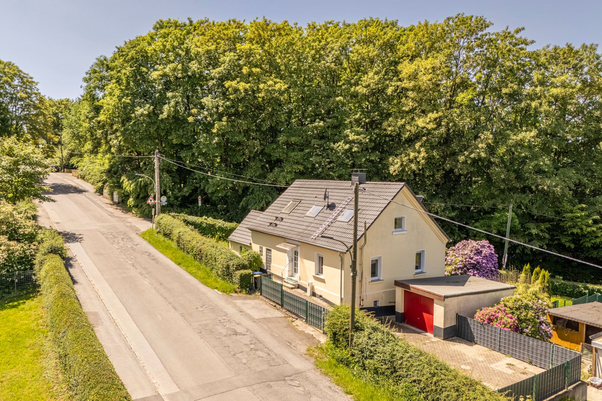 Einfamilienhaus mit dunklem Dach und Garage an einer ruhigen Straße, umgeben von Bäumen.