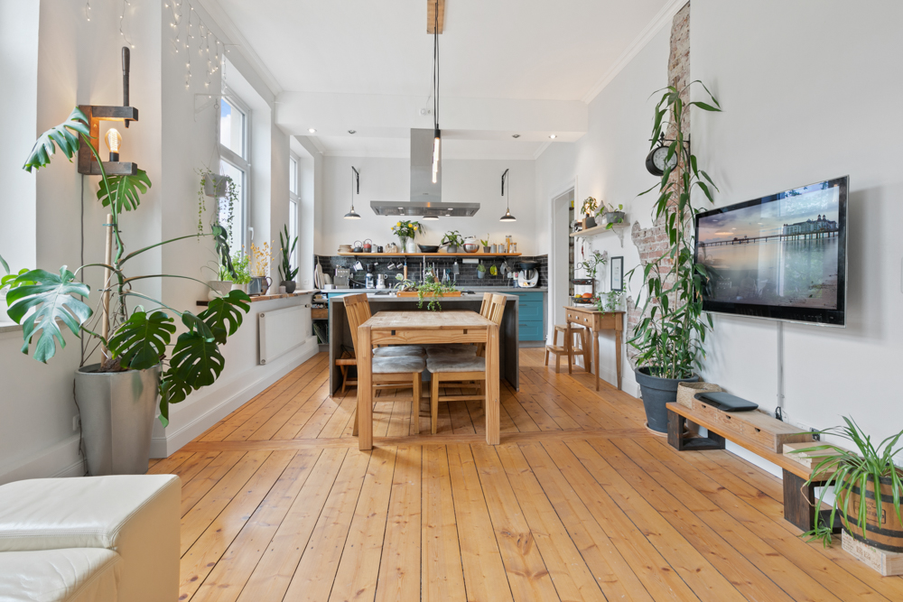 Heller Wohnraum mit Holzboden, Esstisch, offener Küche, vielen Pflanzen und Wandfernseher.