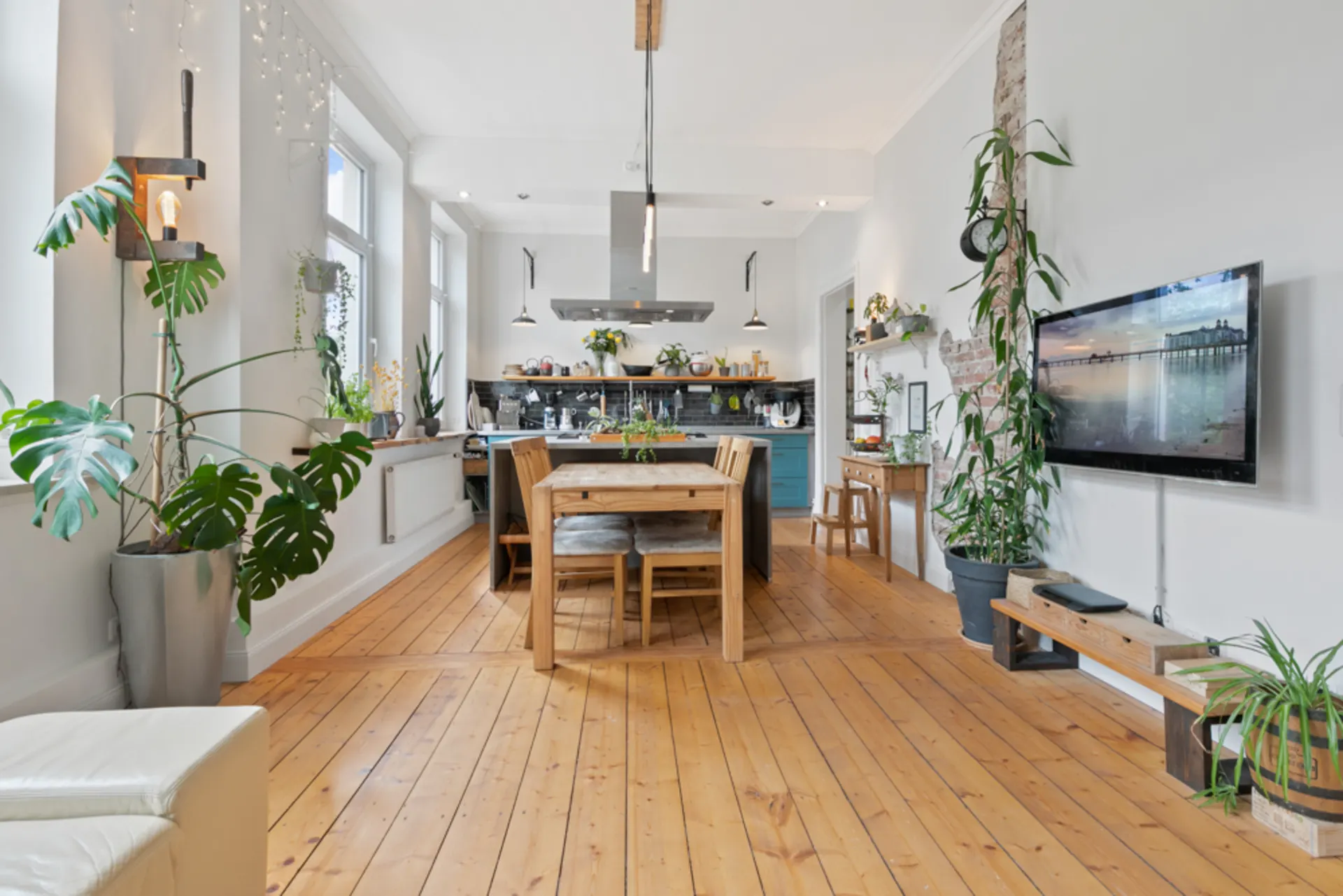 Heller Wohnraum mit Holzboden, Esstisch, offener Küche, vielen Pflanzen und Wandfernseher.