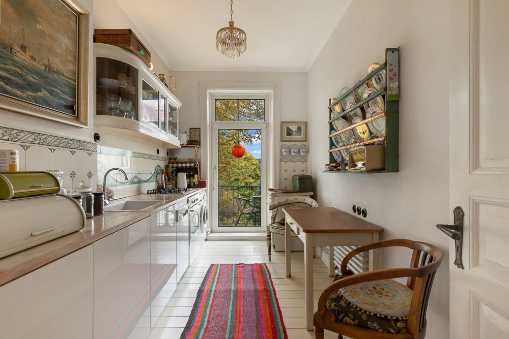 Helle Küche mit Teppichläufer, Tisch, Stuhl und Zugang zu einem Balkon mit Ausblick auf Bäume.