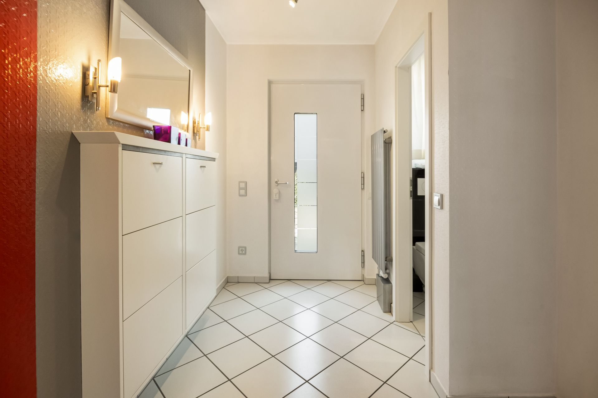 Heller Flur mit weißer Eingangstür, Fliesenboden, Schuhschrank und Spiegel.
