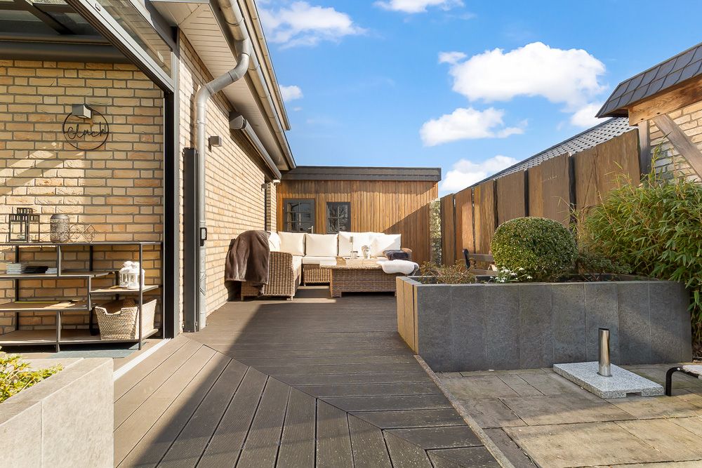 Moderne Terrasse mit Sofa, Holzboden und gepflegten Pflanzen an einem Wohnhaus bei Sonnenschein.