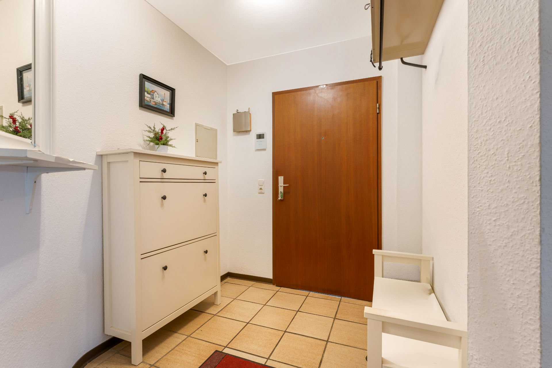 Heller Flur mit Holz-Eingangstür, Kommode, kleiner Bank und Fliesenboden.
