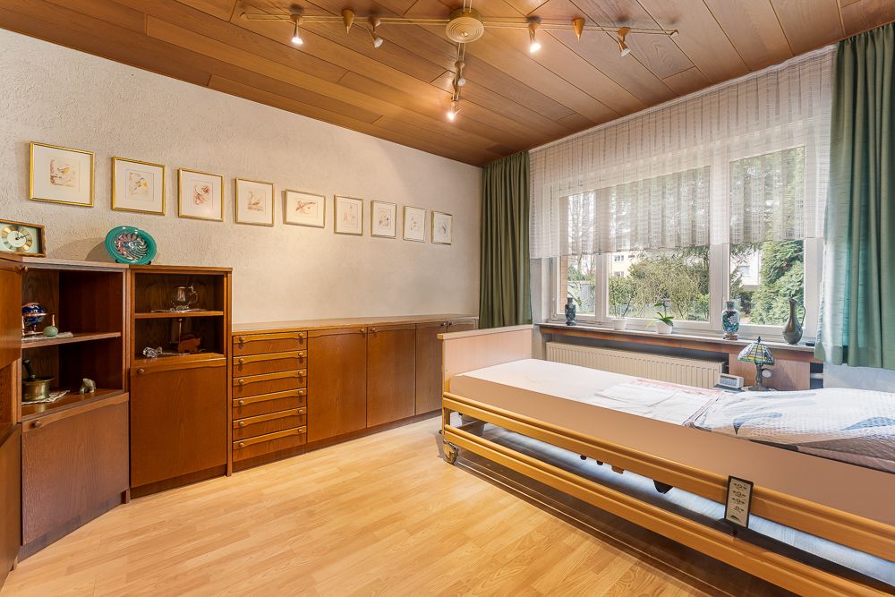 Helles Zimmer mit Holzdecke, Pflegebett, Holzschränken und großem Fenster mit grünen Vorhängen.